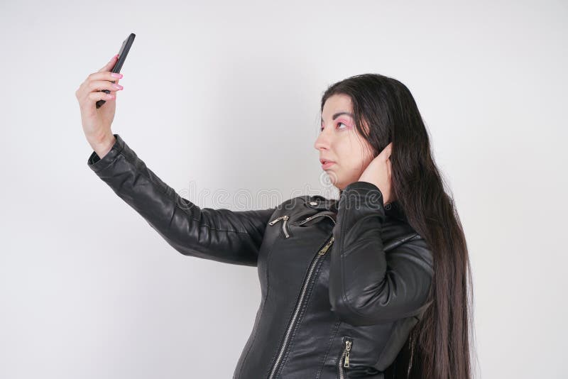 Incantare castana in bomber nero nei supporti di stile della roccia con uno smartphone su un fondo bianco nello studio fotografia stock libera da diritti