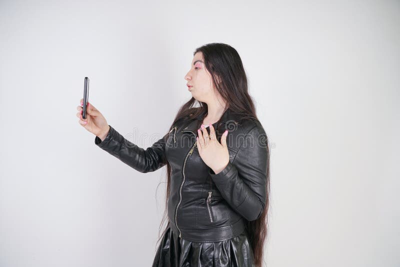 Incantare castana in bomber nero nei supporti di stile della roccia con uno smartphone su un fondo bianco nello studio immagini stock