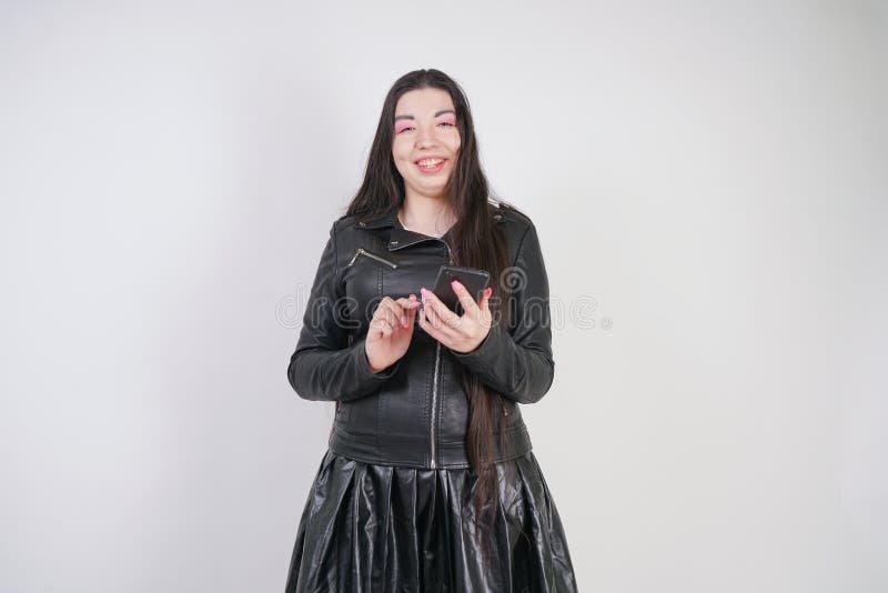 Incantare castana in bomber nero nei supporti di stile della roccia con uno smartphone su un fondo bianco nello studio fotografie stock