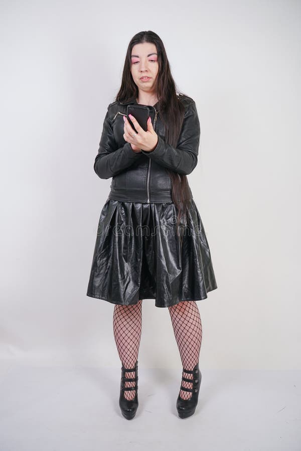 Incantare castana in bomber nero nei supporti di stile della roccia con uno smartphone su un fondo bianco nello studio immagine stock libera da diritti