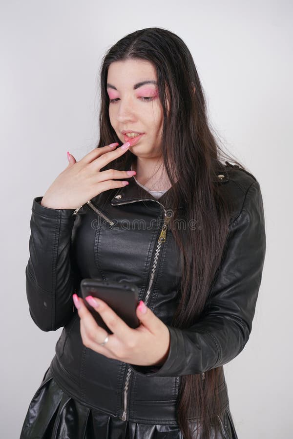 Incantare castana in bomber nero nei supporti di stile della roccia con uno smartphone su un fondo bianco nello studio immagine stock