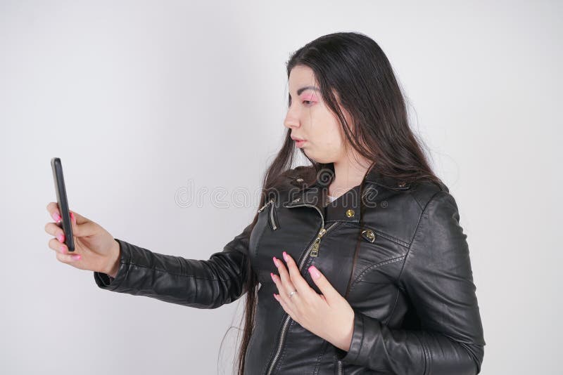 Incantare castana in bomber nero nei supporti di stile della roccia con uno smartphone su un fondo bianco nello studio immagine stock