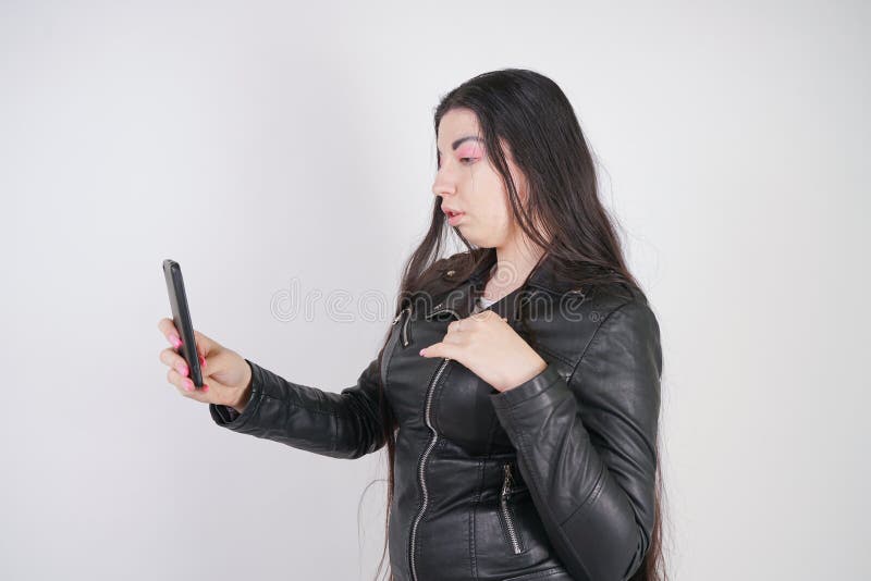 Incantare castana in bomber nero nei supporti di stile della roccia con uno smartphone su un fondo bianco nello studio fotografia stock