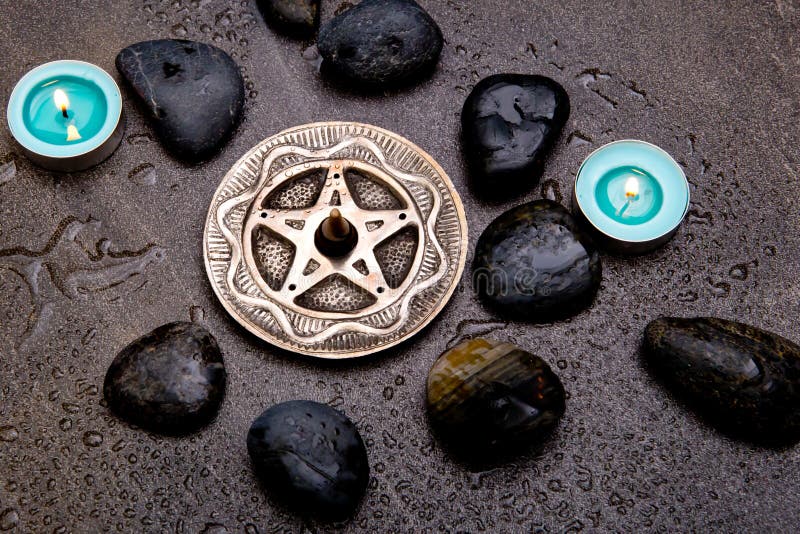 Incense burning in silver pentagram with blue candles and black rocks on gray slate background with water droplets