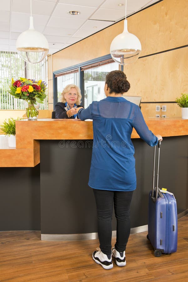 Young woman, with a carry on suitcase next to her, handing over her passport, registering, during checkin at a hotel, where a senior receptionist provides customer service. Young woman, with a carry on suitcase next to her, handing over her passport, registering, during checkin at a hotel, where a senior receptionist provides customer service
