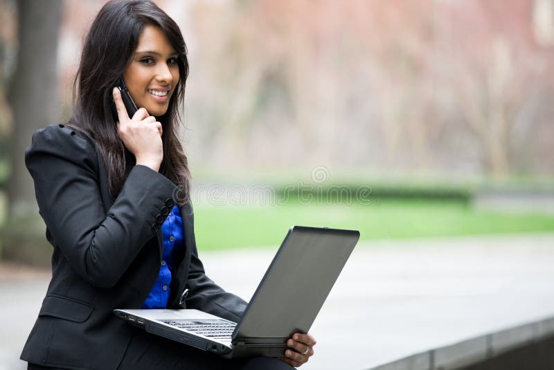A shot of an indian businesswoman talking on the phone and working on her laptop outdoor. A shot of an indian businesswoman talking on the phone and working on her laptop outdoor