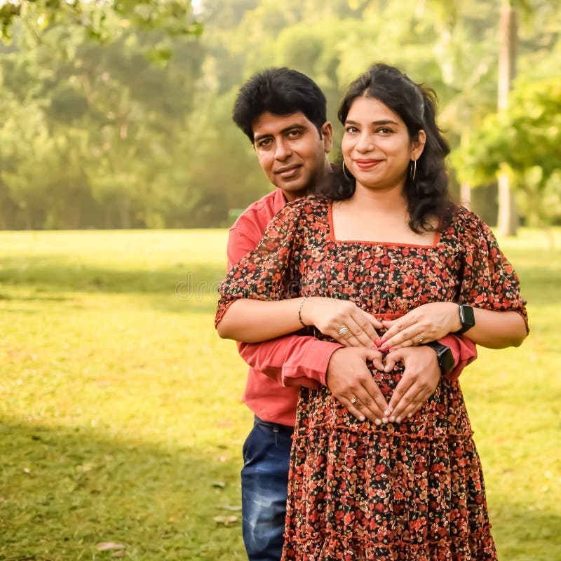 Indian Couple Posing For Maternity Baby Shoot. The Couple Is Posing In A  Lawn With Green Grass And The Woman Is Falunting Her Baby Bump In Lodhi  Garden In New Delhi, India
