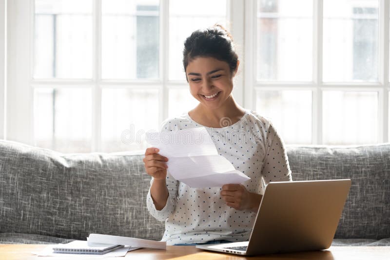 Happy woman holding paper reading good news college admission concept. Indian ethnicity woman sitting on couch at home reading paper notice receive good news stock images