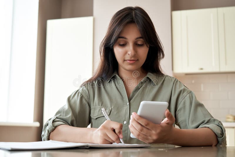 Indian girl holding phone online learn in mobile app write notes at home office.