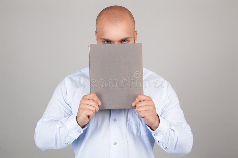 Individuo joven que sostiene un libro delante de su cara mientras que lleva una camisa azul, colocándose en el fondo blanco del e fotos de archivo Individuo joven que sostiene un libro delante de su cara mientras que lleva una camisa azul, colocándose en el fondo blanco del e fotos de archivo