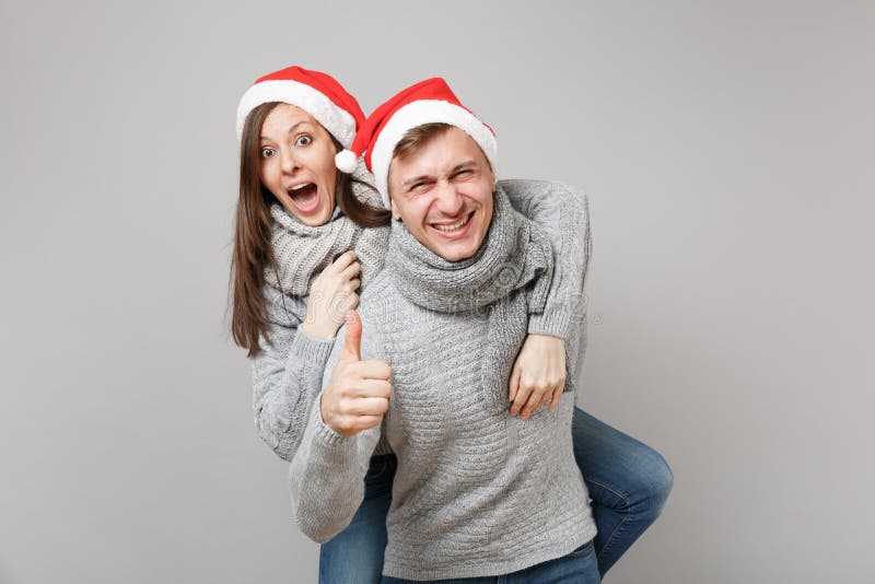 Indivíduo alegre da menina dos pares do divertimento nos scarves cinzentos das camisetas do chapéu vermelho de Santa Christmas is imagem de stock Indivíduo alegre da menina dos pares do divertimento nos scarves cinzentos das camisetas do chapéu vermelho de Santa Christmas is imagem de stock