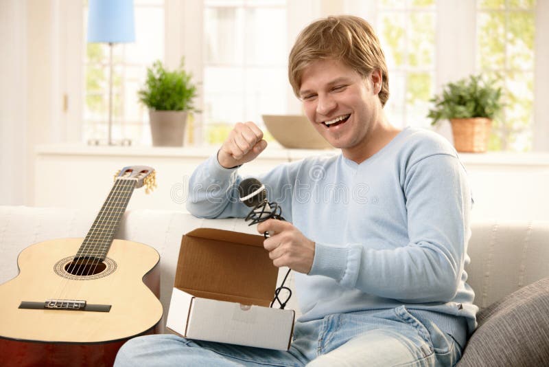 Happy guy getting present, opening box of microphone, sitting on couch with guitar. Happy guy getting present, opening box of microphone, sitting on couch with guitar.