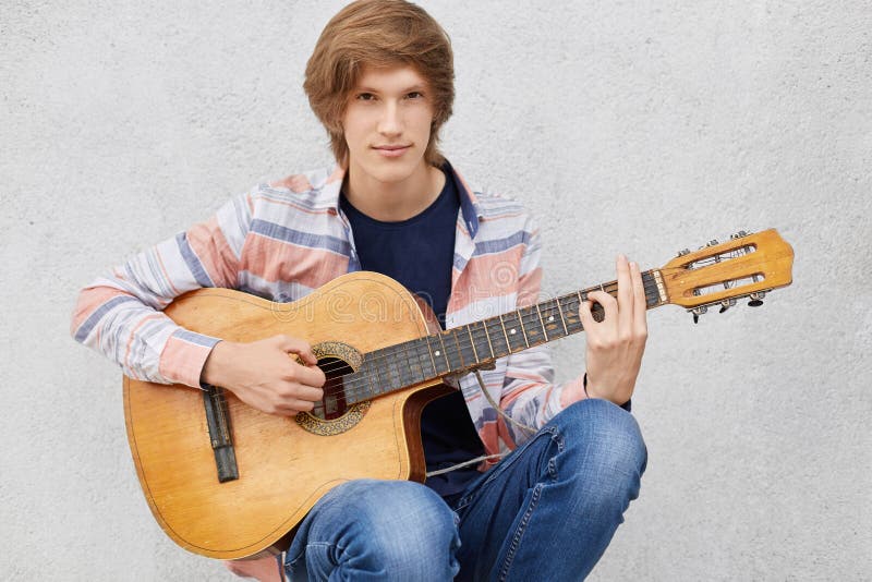 Indivíduo novo considerável com a camisa vestindo e as calças de brim do corte de cabelo à moda que jogam a guitarra, músicas do fotografia de stock Indivíduo novo considerável com a camisa vestindo e as calças de brim do corte de cabelo à moda que jogam a guitarra, músicas do fotografia de stock