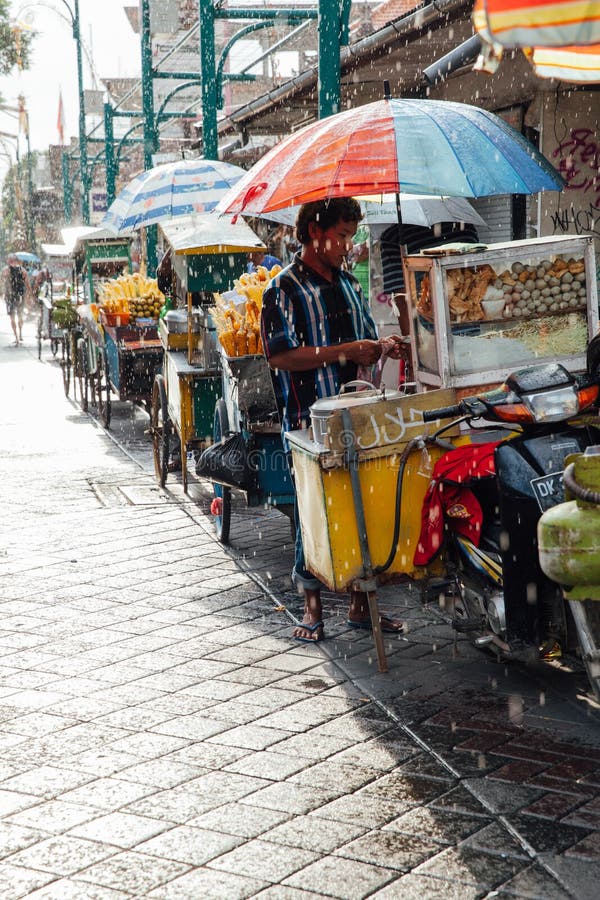 Ubud, Indonesia - March 08, 2016: Indonesian food vendor hides from the rain under umbrella of his stall on the street of Ubud, Bali, Indonesia on March 08, 2016. Ubud, Indonesia - March 08, 2016: Indonesian food vendor hides from the rain under umbrella of his stall on the street of Ubud, Bali, Indonesia on March 08, 2016