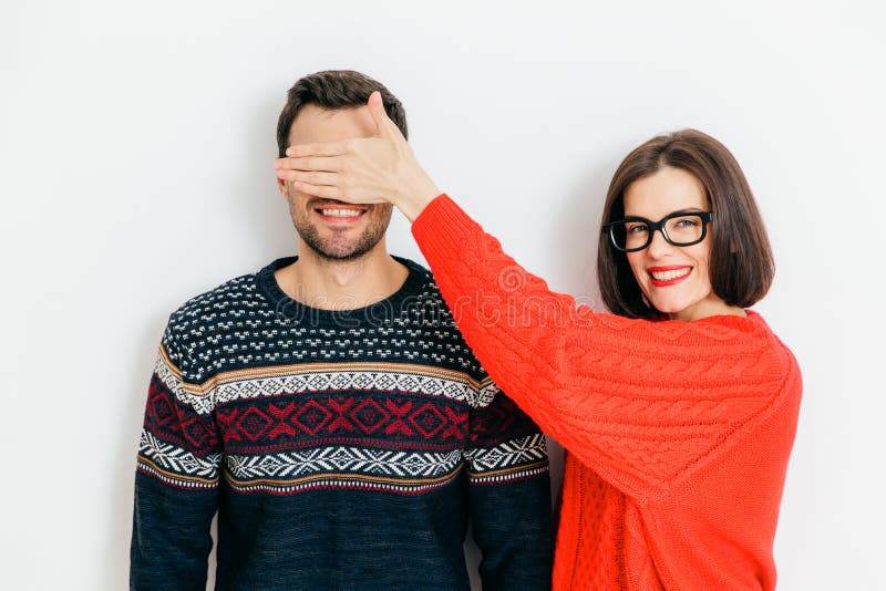 Indoor shot of attractive brunette female with positive expression, covers man`s eyes, wear winter sweaters, stand together again royalty free stock images Indoor shot of attractive brunette female with positive expression, covers man`s eyes, wear winter sweaters, stand together again royalty free stock images