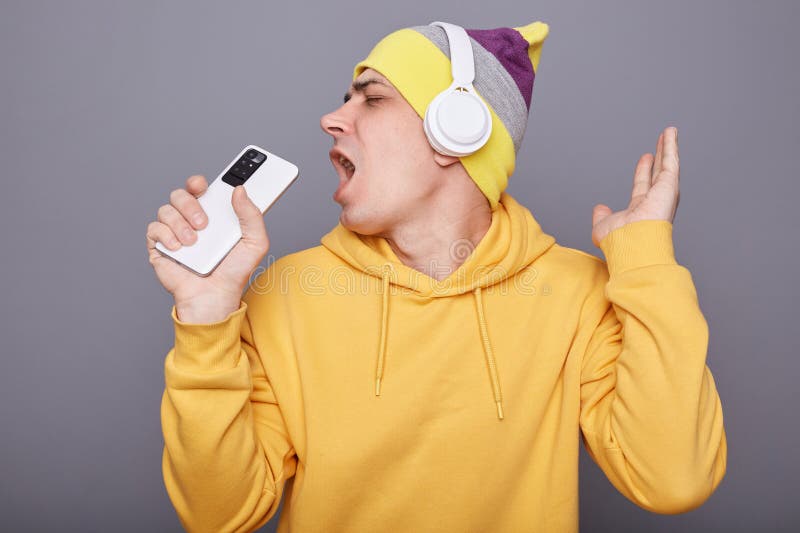 Indoor shot di un allegro uomo soddisfatto con una canzone gialla che canta e utilizza uno smartphone come microfono che si divert fotografie stock