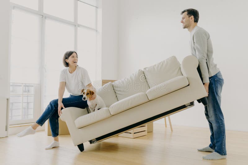 Indoor shot of funny family couple furnish their first home, carry big white sofa, woman teases domestic animal, pose in living