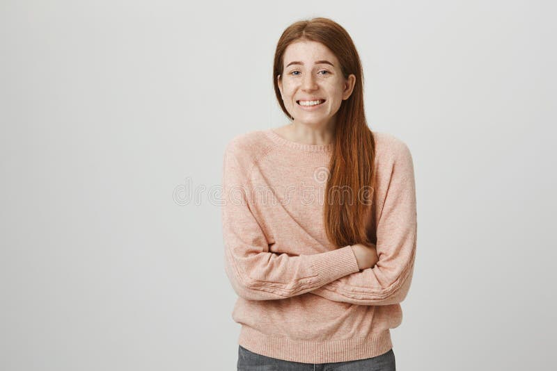 Indoor shot of kind charming ginger girl standing crossed hands as if she is freezing, smiling awkwardly and looking at camera over gray background. Woman went outdoor without wearing jacket.