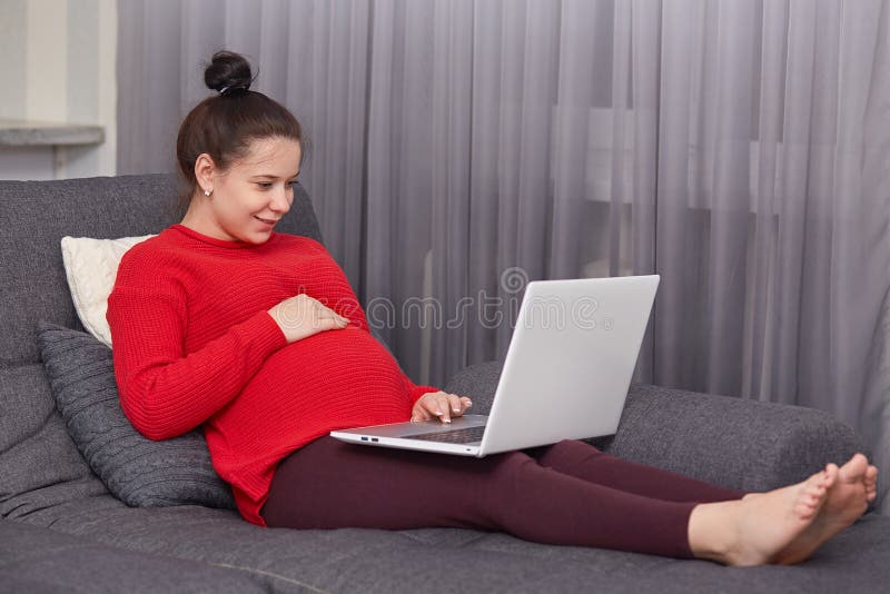 Indoor shot of pleased dark haired pregnant young woman enjoys domestic atmosphhere, keyborads information on laptop computer, stock photography Indoor shot of pleased dark haired pregnant young woman enjoys domestic atmosphhere, keyborads information on laptop computer, stock photography