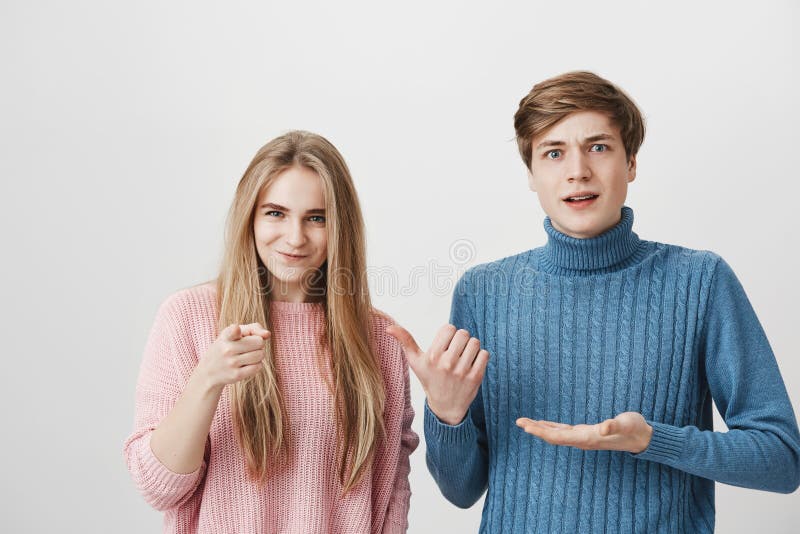 Indoor shot of shocked male and frowning female wearing colourful sweaters looking at camera. Boy pointing with fingers