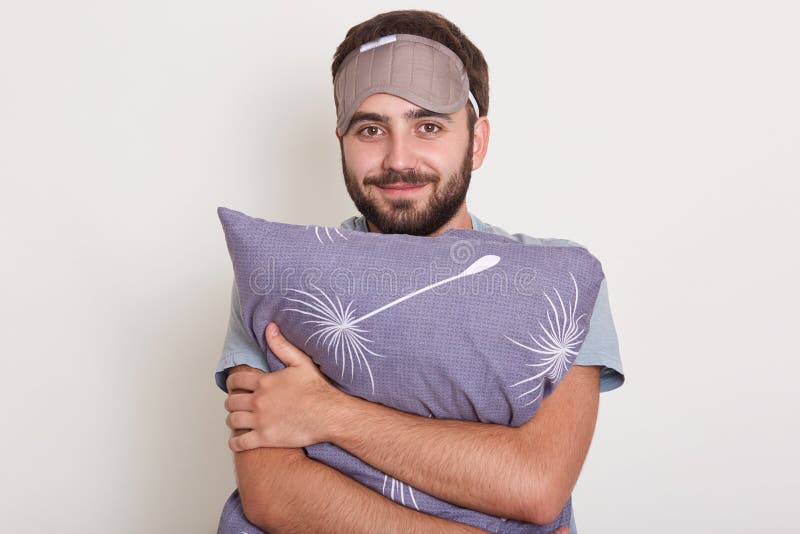 Indoor shot of young handsome man stands smiling and hugging pillow, male posing in bedroom with blindfold on forehead, looking stock photos Indoor shot of young handsome man stands smiling and hugging pillow, male posing in bedroom with blindfold on forehead, looking stock photos