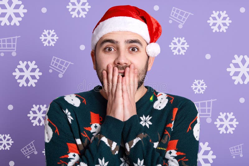 Indoor studio shot of surprised emotional man wearing green sweater with snowmen and red santa claus hat, covering mouth with both royalty free stock photography Indoor studio shot of surprised emotional man wearing green sweater with snowmen and red santa claus hat, covering mouth with both royalty free stock photography