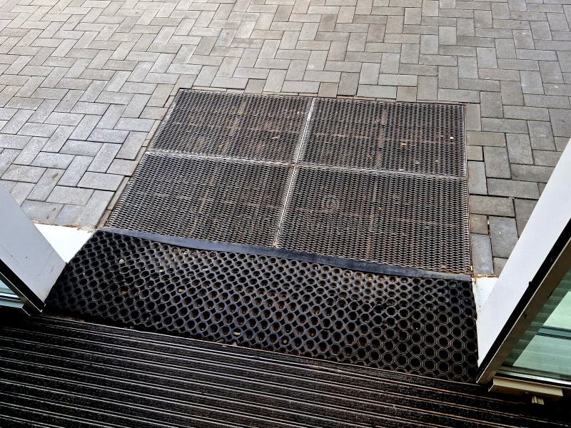 industrial mat cleaning zones at the entrance to the building. A black plastic-metal mat in the shape of an arch or half-circle lies on the limestone mosaic tiles, metal grate, store, mesh wire