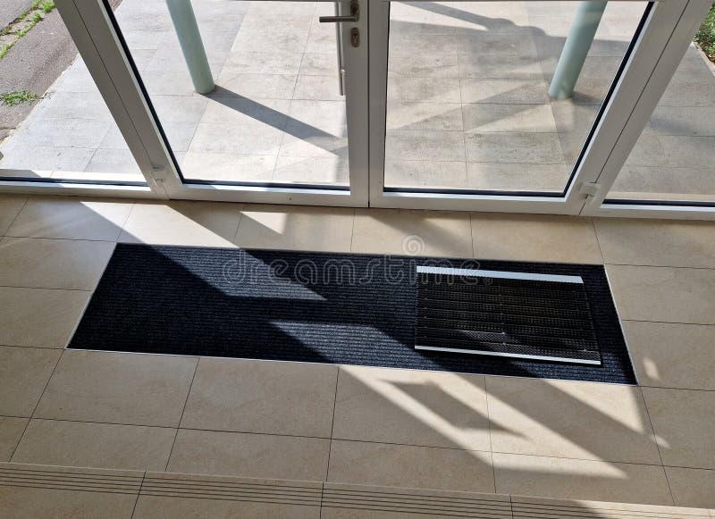 industrial mat cleaning zones at the entrance to the building. A black plastic-metal mat in the shape of an arch or half-circle lies on the limestone mosaic tiles, metal grate, store, mesh wire