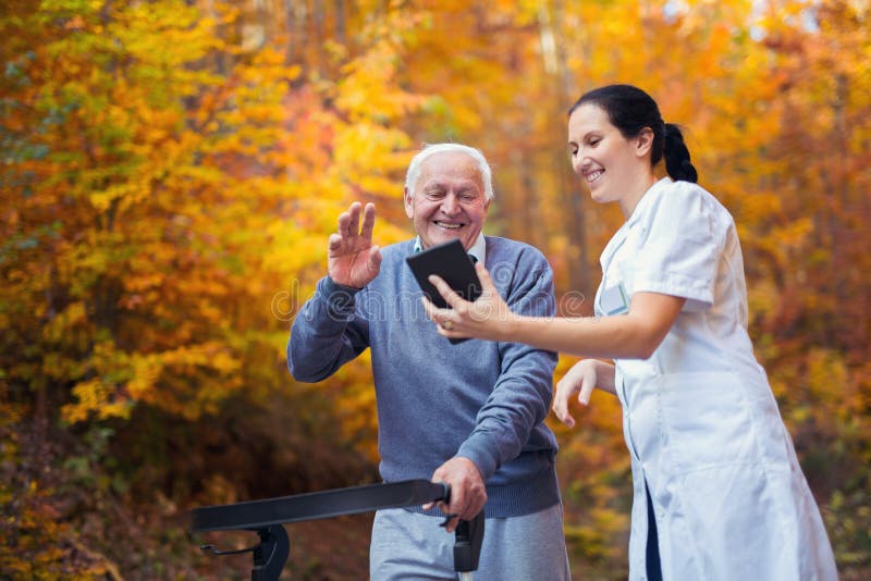 Infirmière et patient supérieur handicapé dans le marcheur à l'aide du comprimé numérique extérieur photos stock Infirmière et patient supérieur handicapé dans le marcheur à l'aide du comprimé numérique extérieur photos stock