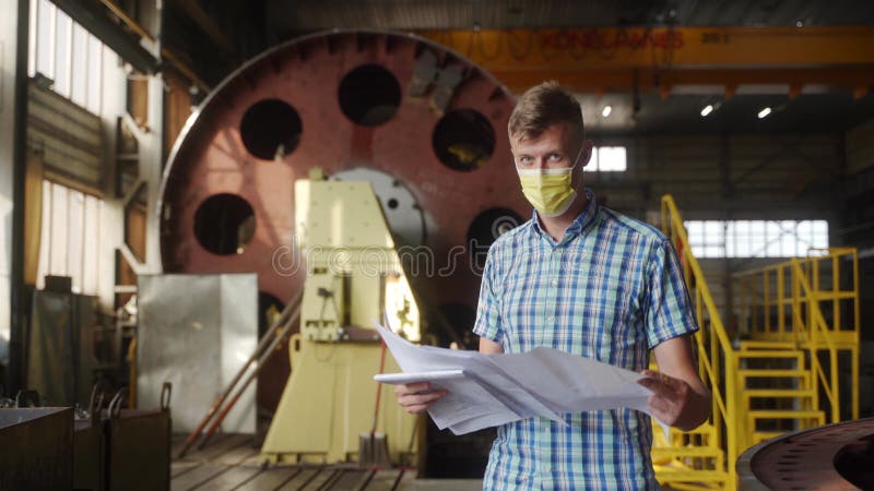 Ingeniero enmascarado examina planes de proyectos en taller industrial. profesional masculino con blueprint evalúa piezas de maqui