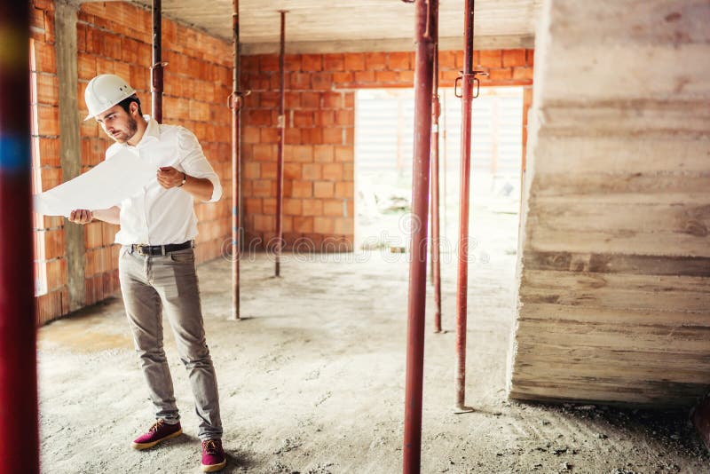 Ingeniero que trabaja en emplazamiento de la obra, dentro de las paredes de ladrillo interiores que llevan el casco y que leen pl fotografía de archivo