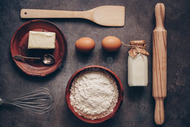 Ingredientes para cocer en un fondo, una harina, una mantequilla, huevos, un rodillo, un batidor y una paleta rústicos oscuros Vi