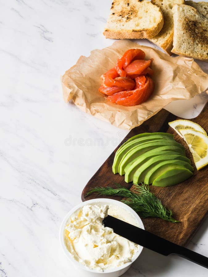 Ingredientes para sanduíches da preparação com queijo creme, salmão, abacate em brindes grelhados no fundo de mármore branco Copi imagem de stock Ingredientes para sanduíches da preparação com queijo creme, salmão, abacate em brindes grelhados no fundo de mármore branco Copi imagem de stock