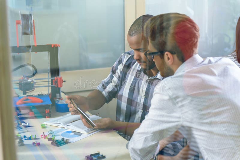 Cheerful male colleagues are entertaining with tablet while 3d printer is working. Men are sitting at desk in office. Cheerful male colleagues are entertaining with tablet while 3d printer is working. Men are sitting at desk in office