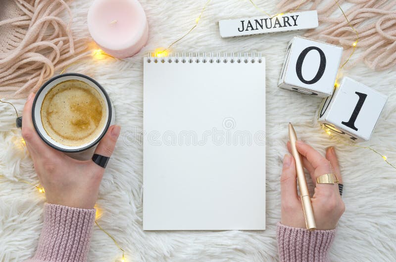 Warm cozy home start new year 1 january on perpetual calendar. Notebook mockup and woman hands holds gold penand cup of coffee, candle, lights. Flat lay top view trendy winter design. Warm cozy home start new year 1 january on perpetual calendar. Notebook mockup and woman hands holds gold penand cup of coffee, candle, lights. Flat lay top view trendy winter design