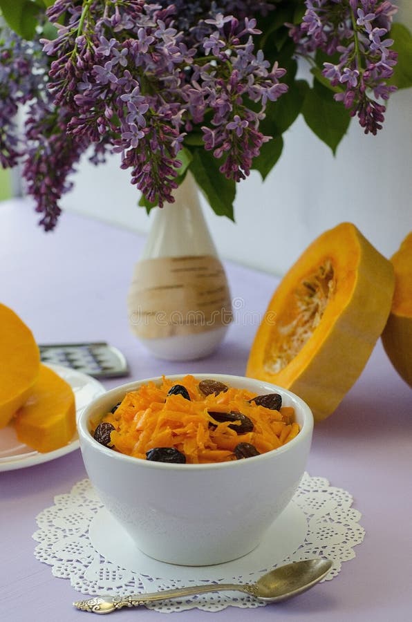 Insalata grattata della zucca con l'uva passa l su una tavola lilla con un mazzo dei fiori lilla Concetto: alimenti crudi, vegani fotografie stock Insalata grattata della zucca con l'uva passa l su una tavola lilla con un mazzo dei fiori lilla Concetto: alimenti crudi, vegani fotografie stock