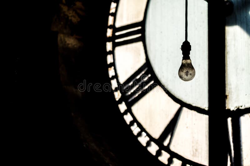 Inside abandoned old ancient clock tower with Roman numerals gears and close up of light bulb hanging down with feathers to illuminate from behind hour minute and second hands ticking