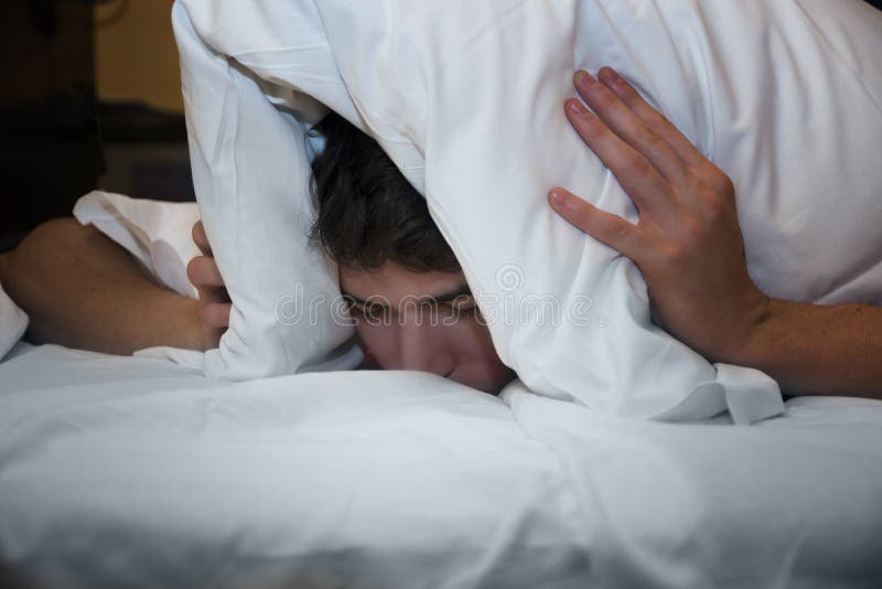 Handsome young male insomniac covering his head and ears trying to block out the sound with a pillow as he is kept awake by loud noises around him. Handsome young male insomniac covering his head and ears trying to block out the sound with a pillow as he is kept awake by loud noises around him