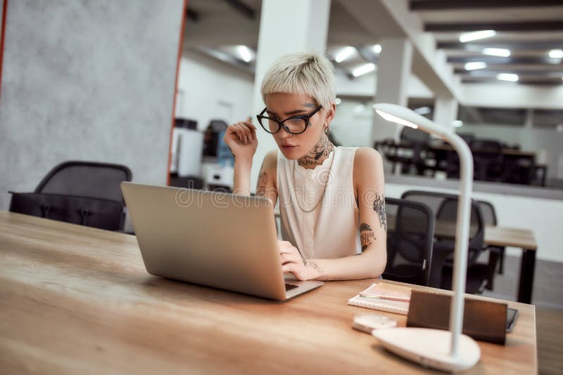 Inspiration. Stylish and beautiful tattooed business lady in eyewear working with laptop while sitting at her desk in