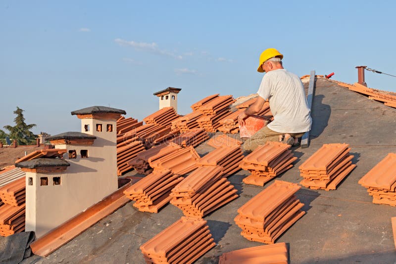 Roofing: construction worker on a roof covering it with tiles - roof renovation: installation of tar paper, new tiles and chimney. Roofing: construction worker on a roof covering it with tiles - roof renovation: installation of tar paper, new tiles and chimney