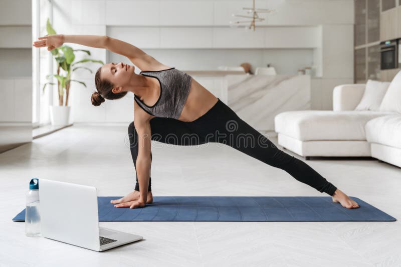 Young attractive woman practicing yoga and stretching body at home using laptop for online class or virtual tutorials. Girl standing in Utthita parsvakonasana exercise, doing Side Angle pose. Wellness. Young attractive woman practicing yoga and stretching body at home using laptop for online class or virtual tutorials. Girl standing in Utthita parsvakonasana exercise, doing Side Angle pose. Wellness