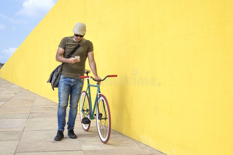 Integral de hombre usando el teléfono elegante mientras que hace una pausa la bicicleta contra la pared amarilla en la acera en c fotos de archivo libres de regalías Integral de hombre usando el teléfono elegante mientras que hace una pausa la bicicleta contra la pared amarilla en la acera en c fotos de archivo libres de regalías