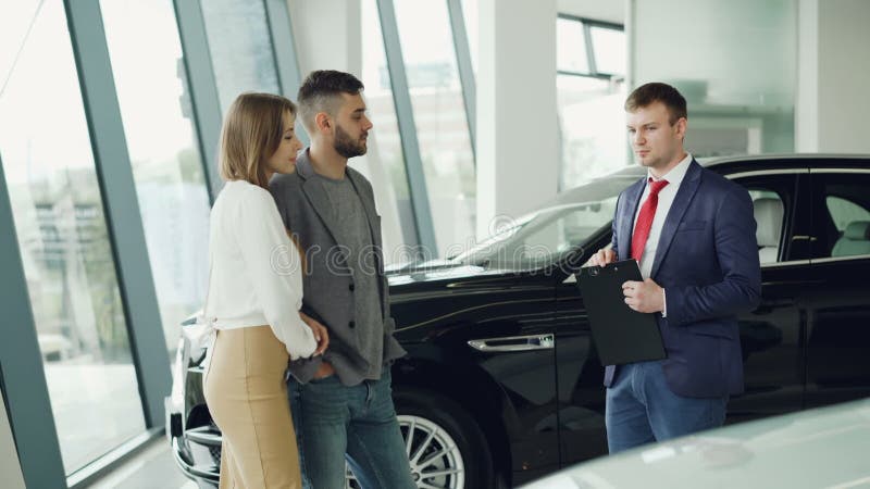 Interested couple man and woman are talking to manager in car dealership while he is telling them about luxury auto