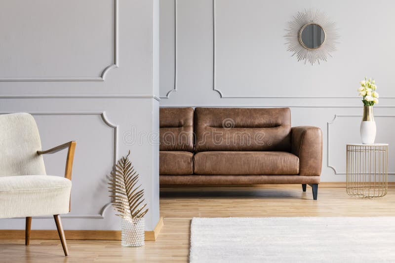 Open space living room interior with decorative mirror on wall with wainscoting, brown leather sofa, fresh roses on end table and gold fern leaf in glass vase placed by white armchair in real photo. Open space living room interior with decorative mirror on wall with wainscoting, brown leather sofa, fresh roses on end table and gold fern leaf in glass vase placed by white armchair in real photo