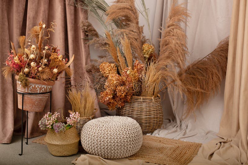 Interior of house is decor in brown tones: wooden chair, knitted pouf, wicker baskets, vases with dried flowers and pampas grass.