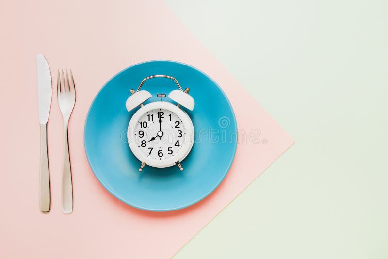 Intermittent fasting concept. White alarm clock on empty blue plate with knife and fork on pink-green background. Eight hour feeding window concept. Top view, copy space. Intermittent fasting concept. White alarm clock on empty blue plate with knife and fork on pink-green background. Eight hour feeding window concept. Top view, copy space