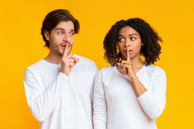 Common secret. Interracial couple holding fingers on lips, making shh sign, hiding some information, yellow studio background with empty space. Common secret. Interracial couple holding fingers on lips, making shh sign, hiding some information, yellow studio background with empty space