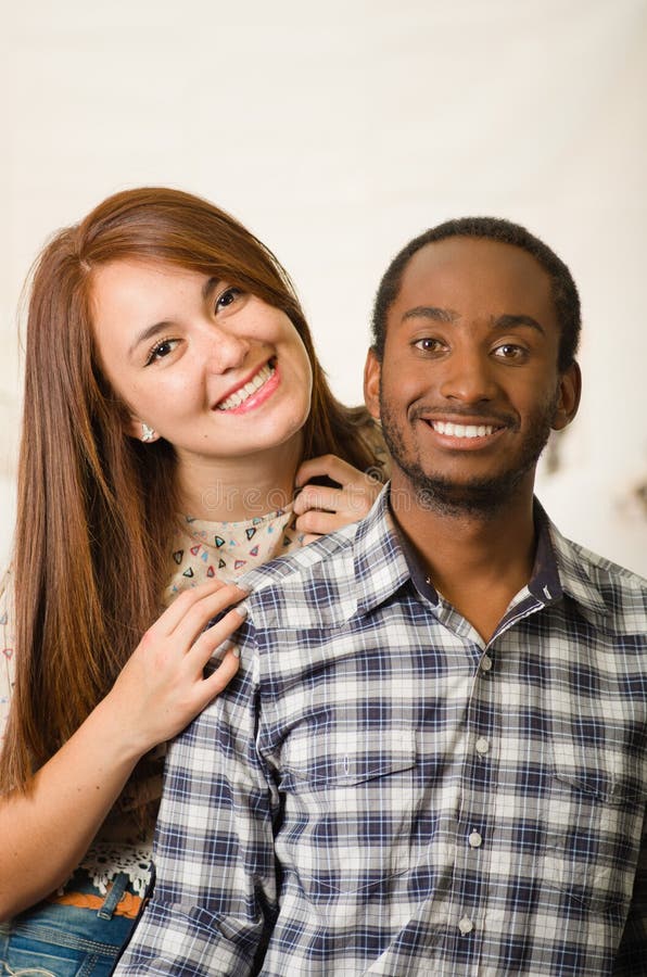 Interracial Charming Couple Wearing Casual Clothes Sitting on Wooden Surface Posing for Camera