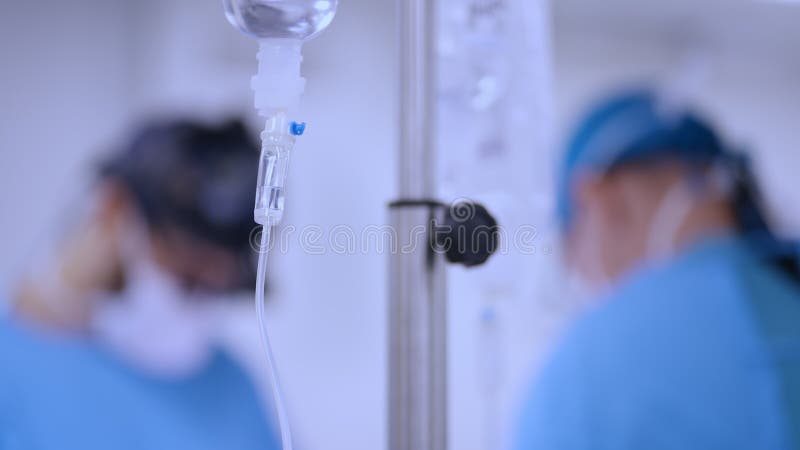 Intravenous drip in focus, surgical team performing surgery in background, out of focus