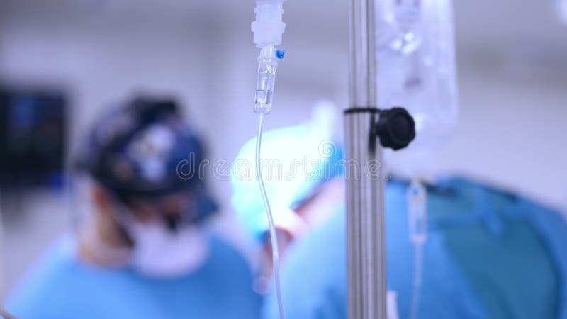 Intravenous drip in focus, surgical team performing surgery in background, out of focus